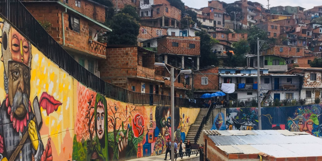 Long mural wall beside a walkway with the brick hillside houses of Comuna 13 rising behind