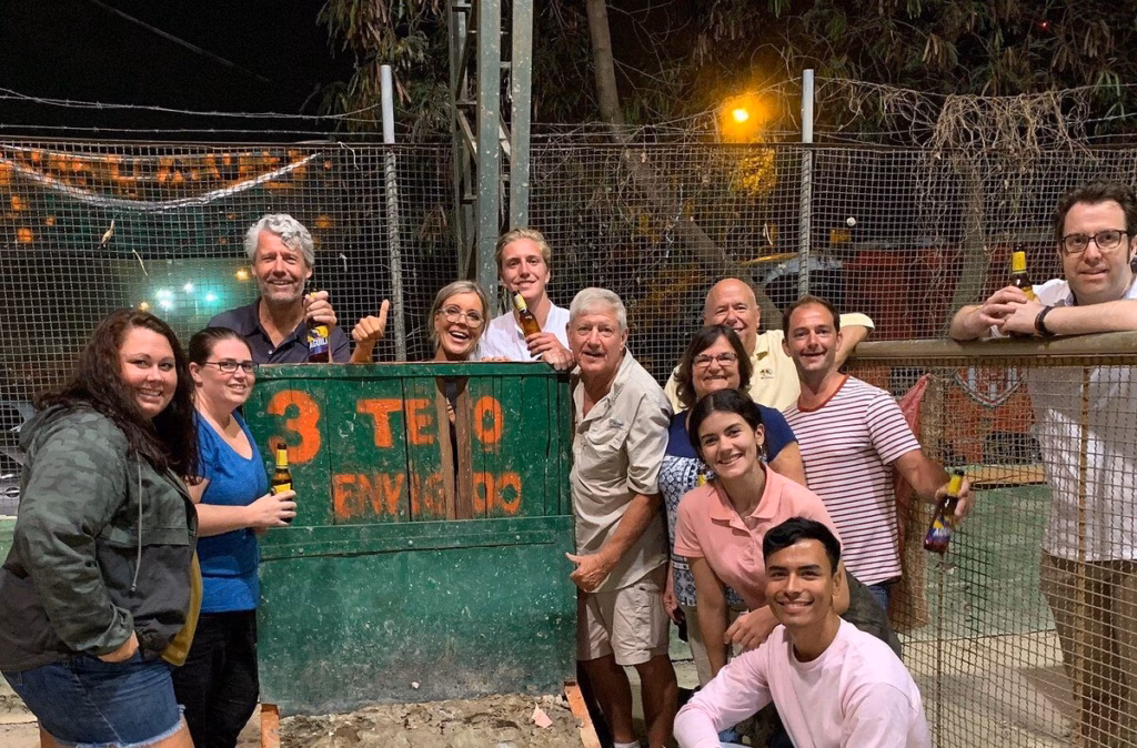 Group posing with beers in front of a green tejo scoreboard behind the court netting