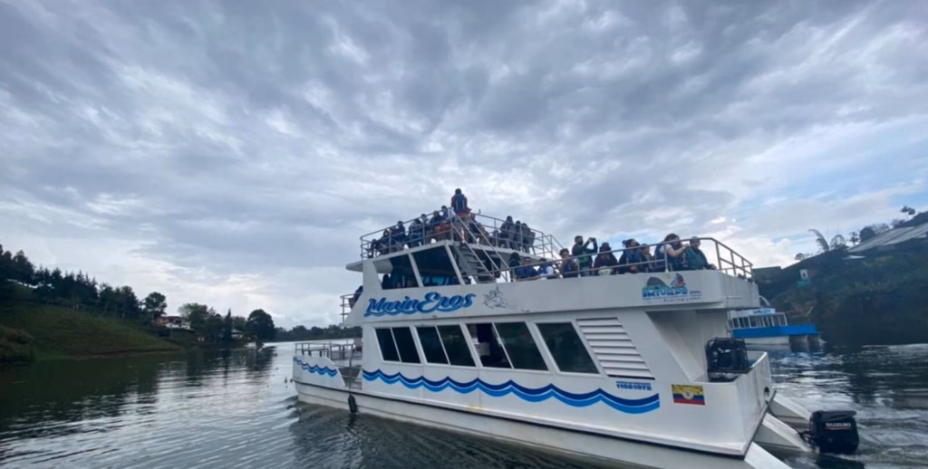 Double-deck tour boat carrying passengers across the Guatapé reservoir under a cloudy sky