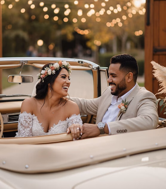 Bride and groom laughing inside a vintage car with warm lights in the background.